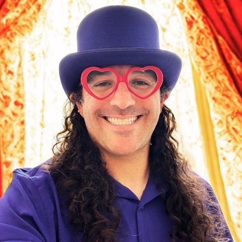 ony Cañas smiling warmly while wearing red heart-shaped glasses, a purple top hat, and a purple collared shirt, standing in front of vibrant gold and red curtains.